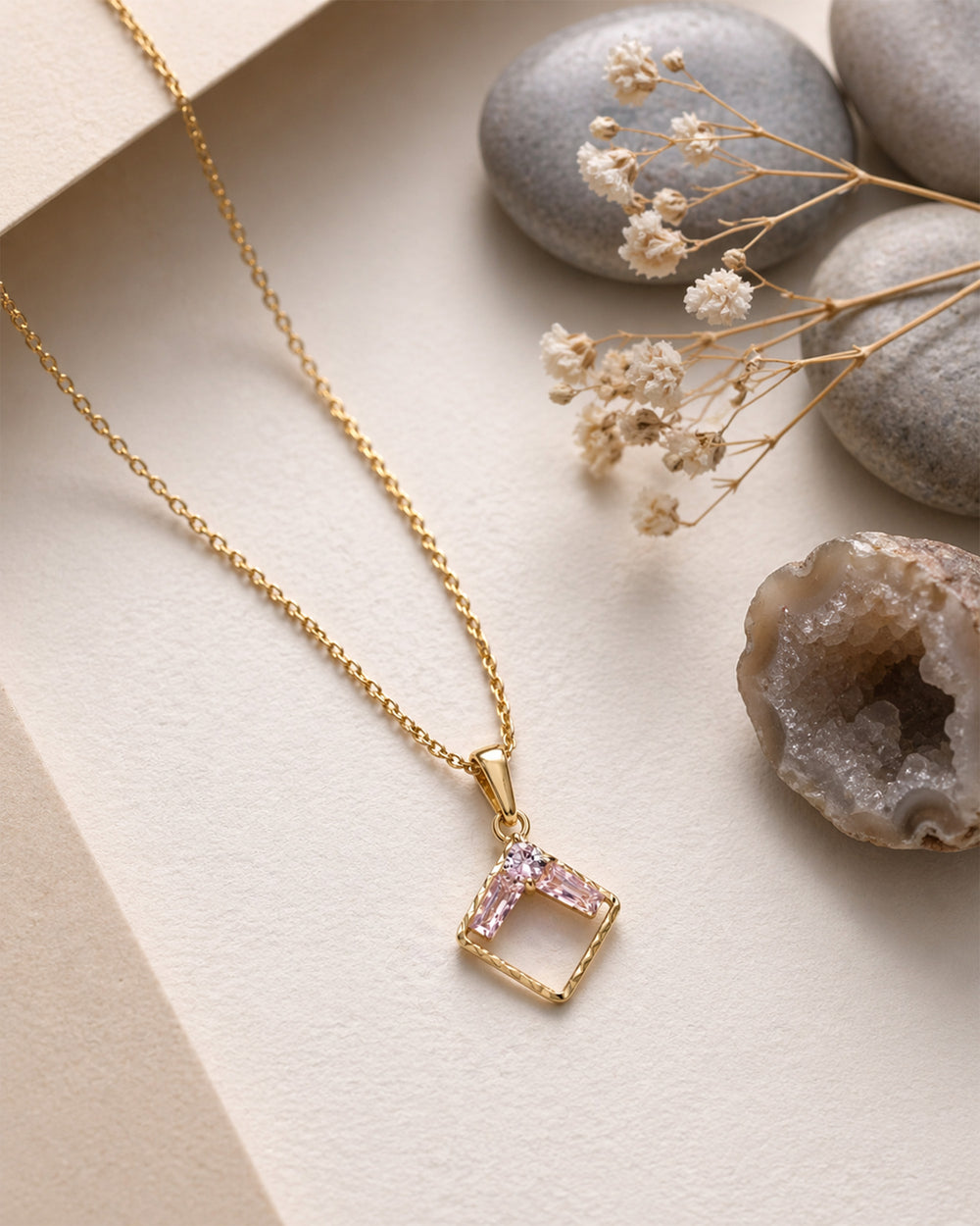 Gold necklace with a square pendant on a neutral background with stones and dried flowers.