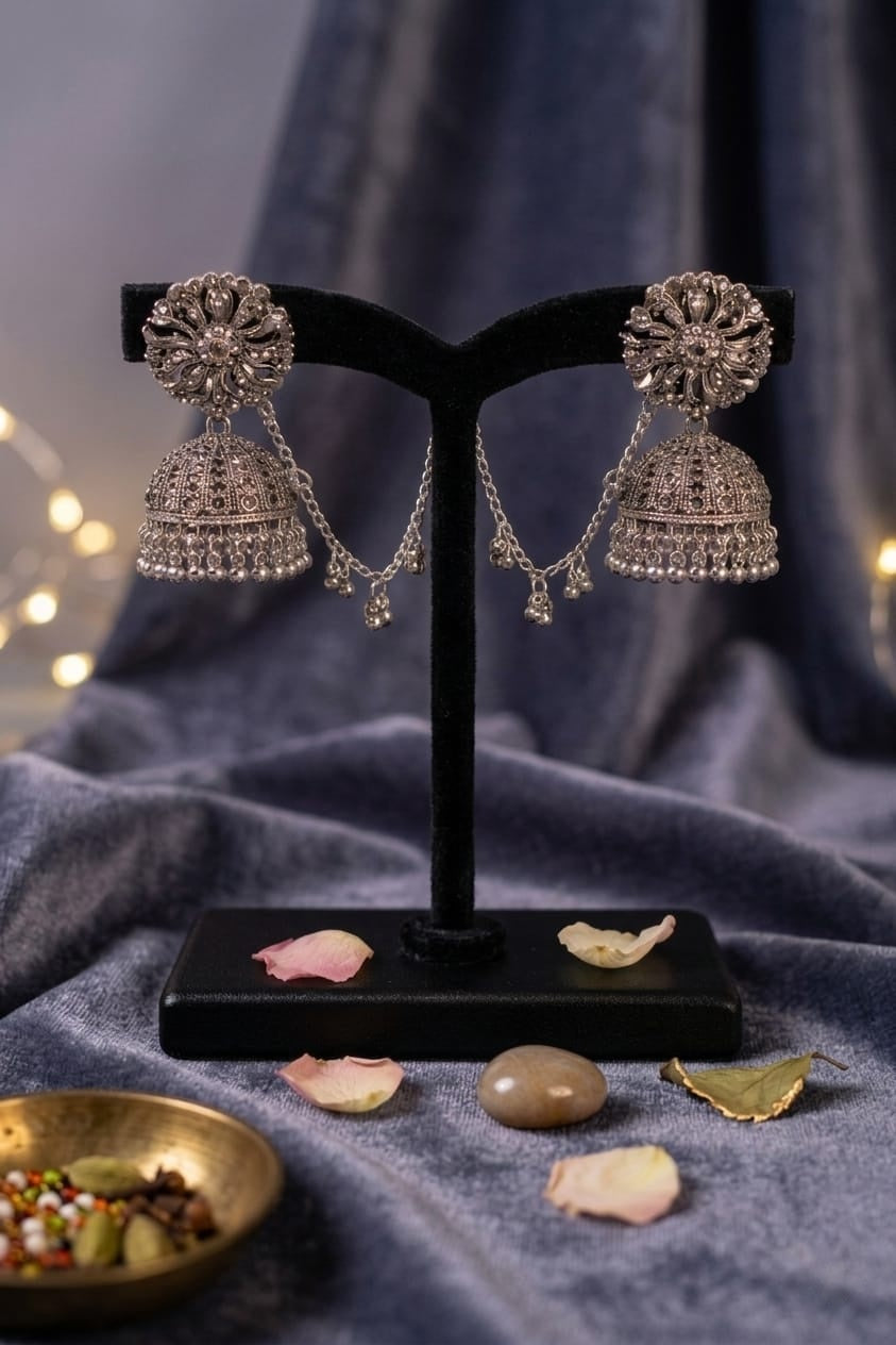 Silver jhumka earrings on a black stand with decorative elements on a blue fabric background.