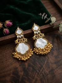 Goldden and white earrings on a wooden surface with green fabric and dried flowers in the background