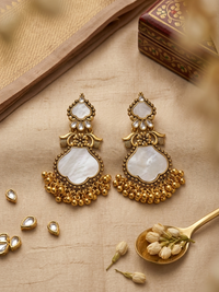 Ornate goldden earrings with white stones on a beige surface with a wooden box in the background.