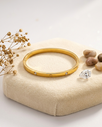 Gold bracelet with small diamonds on a beige cushion with decorative stones and dried flowers.